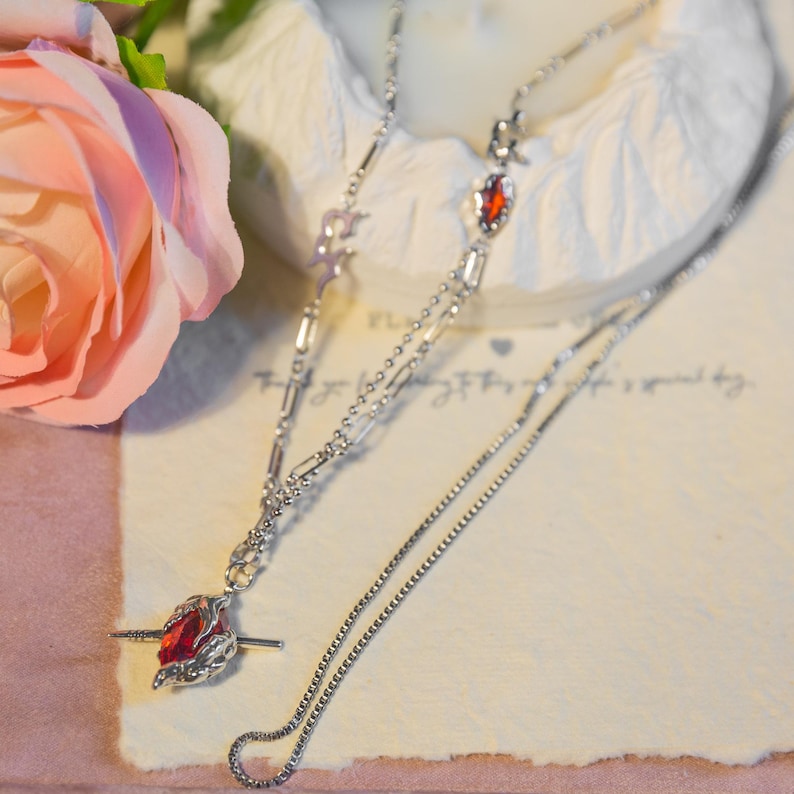May include: A silver necklace with a red gemstone pendant. The pendant is a stylized rose with a red stone at its center. The necklace has a delicate chain design and is displayed on a light-colored surface.