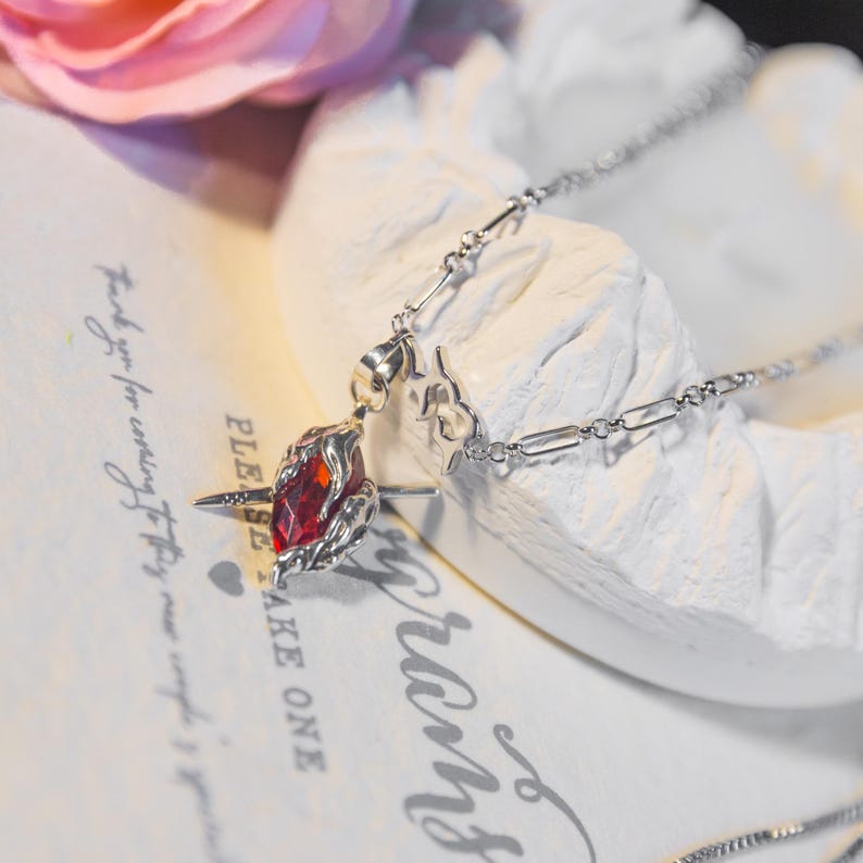 May include: A silver necklace featuring a pendant with a large, faceted red gemstone. The pendant is surrounded by a detailed silver setting. The necklace is displayed on a textured white surface with faint script in the background.
