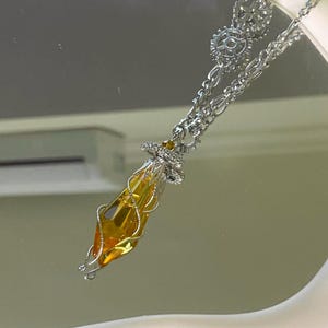May include: A silver-toned necklace featuring a pendant with a faceted, amber-colored crystal encased in a silver wire design. The necklace has a chain with decorative gears and a small, sparkling accent at the top of the pendant.