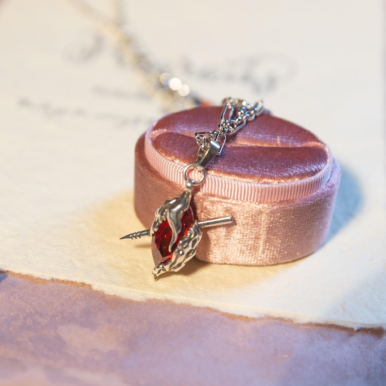 May include: A silver necklace with a pendant shaped like a heart pierced by a silver arrow. The heart is a deep red gemstone. The necklace rests on a pink velvet jewelry box, with a soft, blurred background.