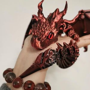 Peut inclure: Un ensemble d'accessoires sur le thème du dragon rouge. L'ensemble comprend une bague tête de dragon, une armure de main en forme de griffes et un bracelet avec des perles rouges. Le dragon a des yeux rouges et des écailles texturées. Les ailes sont visibles en arrière-plan.