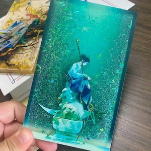 Op de afbeelding: Een rechthoekig harskunstwerk met een figuur in blauwe en witte kleding, zittend op een decoratieve basis. De achtergrond is een levendig groen met bamboe- en glitteraccenten. Het stuk wordt door een hand vastgehouden.