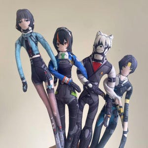 May include: Four handmade fabric dolls with anime-style designs. Each doll features a unique outfit and hairstyle, with detailed facial features. The dolls are primarily in shades of blue, black, and gray, with accents of orange and white. The dolls are standing close together.