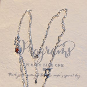 May include: A silver necklace with a square, reddish-brown gemstone and decorative flame-like charms. The necklace has a delicate chain design and is displayed on a cream-colored surface with the words "Please Take One".