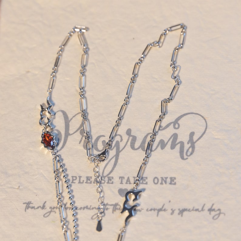 May include: A silver necklace with a square, reddish-brown gemstone and decorative flame-like charms. The necklace has a delicate chain design and is displayed on a cream-colored surface with the words "Please Take One".