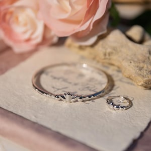 Könnte beinhalten: Ein silbernes Armband- und Ring-Set mit floralem Design. Das Armband ist ein dünnes Band mit einem sich wiederholenden Muster aus stilisierten Blumen. Der Ring hat ein ähnliches Design. Der Schmuck liegt auf Papier mit Schrift, neben einer rosa Rose.