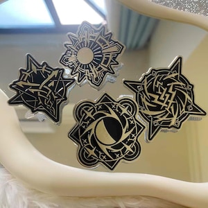 May include: Four black and gold metal pins with intricate geometric designs. The pins are in various star and sunburst shapes, with detailed patterns and a metallic finish. The pins are displayed on a reflective surface.