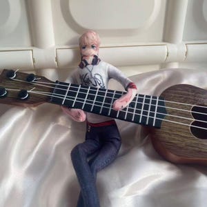 May include: A doll figure with a ukulele. The doll is wearing a white long-sleeve shirt with a graphic design, blue jeans, and headphones. The ukulele is brown with black frets and strings. The doll is holding the ukulele.