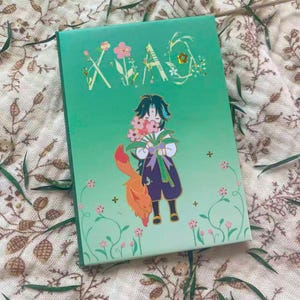 Peut inclure: Un carnet vert avec le mot "XIAO" en lettrage floral. La couverture présente un personnage de dessin animé tenant des fleurs, avec un renard orange. Des fleurs roses et des éléments décoratifs entourent le personnage. Le carnet est sur un tissu à motifs.