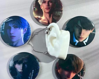 Op de afbeelding: Een verzameling van vijf ronde buttonspins met portretten van anime-stijl personages. Een wit, oorvormig displaystuk met zilveren oormanchetten en een ketting staat in het midden. De buttons hebben een glanzende afwerking.