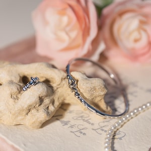 Könnte beinhalten: Ein silbernes Armband und ein passender Ring mit dekorativen Details. Der Schmuck wird auf einer strukturierten, hellen Oberfläche präsentiert, mit einer Perlenkette und zwei rosa Rosen im Hintergrund. Das Armband hat einen Durchmesser von etwa 6,35 cm.