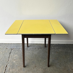 May include: A vintage yellow drop-leaf table with dark brown legs. The table has a rectangular shape with two drop leaves that can be folded down. The table is approximately 30 inches long and 24 inches wide when fully extended.