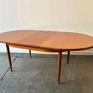 May include: Oval wooden dining table with a light brown finish. The table has four tapered legs and a smooth surface. The table is extended, showing the additional leaf. A classic piece of furniture.