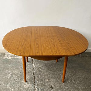 May include: A mid-century modern oval dining table with a warm wood grain finish. The table has four slender, tapered legs in a contrasting orange-brown hue. The table appears to have a leaf extension for added surface area.