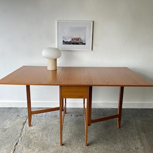 May include: A rectangular wooden drop-leaf table with a warm brown finish and a central drawer, featuring two hinged side panels that extend the surface. A mushroom-shaped table lamp with a white glass shade and a beige base sits on the table.