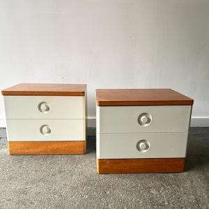 Könnte beinhalten: Zwei passende Nachttische im Mid-Century-Modern-Design. Jeder Nachttisch hat eine helle Holzplatte, zwei weiße Schubladen mit runden Griffen und einen hellen Holzsockel. Sie stehen vor einer weißen Wand.