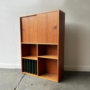 May include: A mid-century modern wooden cabinet with a warm, natural finish. The cabinet features a sliding door on top and open shelving below, including a section with vertical dividers. The design is simple and functional, suitable for storage.