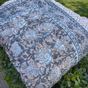 Peut inclure: Une courtepointe à motifs floraux dans les tons pêche, blanc, gris et beige. La courtepointe présente un grand motif floral avec une bordure de motifs géométriques gris et beiges. La courtepointe est pliée et posée sur des feuilles vertes.
