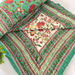 May include: A folded green and red floral quilt with a matching rolled-up quilt. The quilt features a repeating floral pattern in red and white on a green background. The quilt has a white centre with red and green floral designs.