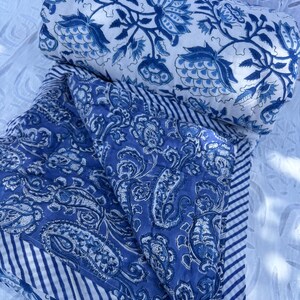 Peut inclure: Une courtepointe bleue et blanche pliée avec un motif floral et un oreiller cylindrique assorti. La courtepointe présente un motif cachemire et une bordure rayée. La palette de couleurs est principalement bleue et blanche.