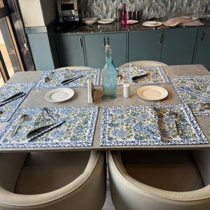 Puede incluir: Una mesa de comedor para seis personas con manteles individuales, servilletas y platos con estampado floral. La cubertería está cuidadosamente dispuesta. Una botella de vidrio azul y un pequeño salero blanco están en el centro.