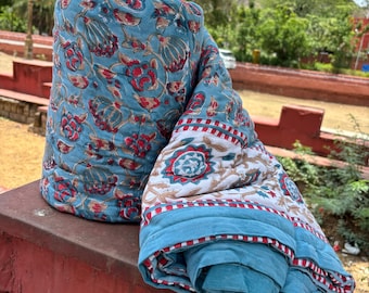 May include: A folded blue quilt with a floral pattern in red and white. The quilt is folded on a red surface, with a portion of the quilt rolled up. The edges of the quilt have a white and red striped border.