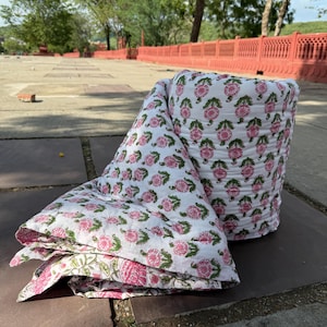 Może przedstawiać: Biała kołdra z powtarzającym się wzorem różowych kwiatowych wzorów i zielonych liści. Kołdra jest częściowo zwinięta i częściowo złożona, prezentując skomplikowany wzór. Pościel jest umieszczona na kamiennej powierzchni.