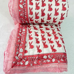 Puede incluir: Un edredón blanco y rojo enrollado con un patrón repetido de conejitos rojos. El edredón tiene un borde a rayas rojo y blanco y un borde floral decorativo en rojo. El edredón está hecho de material suave.