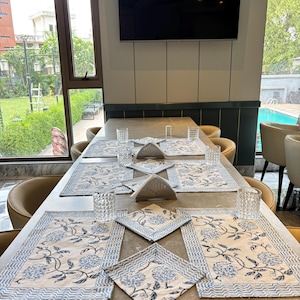 May include: A dining table set with cream-coloured placemats and napkins, adorned with blue floral patterns and a wavy border. Clear, patterned drinking glasses are arranged on the table. The setting is ready for a meal.