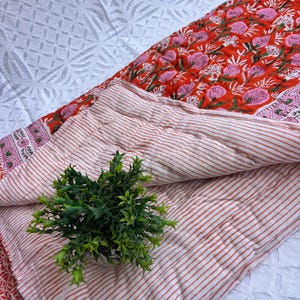 May include: A quilted blanket with a floral pattern in red and pink. The reverse side features a striped pattern in red and white. A small green plant sits on top of the blanket.
