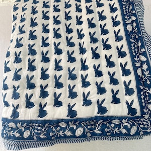 May include: A white and blue quilt with a repeating pattern of navy blue bunny silhouettes. The quilt has a decorative border with floral and leaf designs. The quilt is a square shape.
