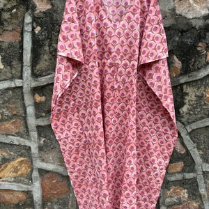 May include: A pink caftan with a V-neck and short sleeves, hanging on a wooden hanger. The fabric features a repeating pattern of brown and pink floral shapes. The garment is made of a lightweight material.
