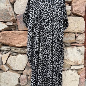 Könnte beinhalten: Ein schwarz-weiß gemustertes Kaftankleid hängt an einem Holzbügel. Das Kleid hat ein sich wiederholendes Muster aus unregelmäßigen Formen. Das Kleidungsstück wird vor dem Hintergrund einer Steinmauer präsentiert.