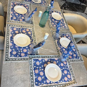 Może przedstawiać: Stół jadalny z sześcioma nakryciami. Każde nakrycie zawiera biały talerz, niebieską podkładkę w kwiaty, złożoną serwetkę i szklankę wody. Zielona szklana butelka oraz solniczka i pieprzniczka znajdują się na środku.