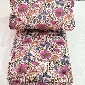 Op de afbeelding: Een kussen en gevouwen stof met bloemenpatroon. Het ontwerp toont grote magenta bloemen, blauwe knoppen en bruine bladeren op een witte achtergrond. De stof is waarschijnlijk katoen of een vergelijkbaar materiaal, geschikt voor beddengoed of woondecoratie.