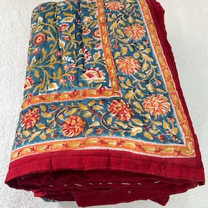 Könnte beinhalten: Eine gefaltete Steppdecke mit tealfarbenem Hintergrund und einem Blumenmuster in Orange, Gelb und Weiß. Die Steppdecke hat einen roten Rand und besteht aus einem weichen, gesteppten Stoff. Das Blumenmuster wiederholt sich.