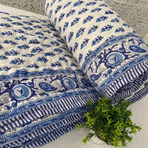 Könnte beinhalten: Eine weiß-blaue Steppdecke mit floralem Muster. Die Steppdecke ist gefaltet und gerollt und zeigt ein detailliertes Bordürendesign. Das blaue Blumenmuster wiederholt sich auf der Oberfläche der Steppdecke. Die Steppdecke besteht aus weichem, gestepptem Material.