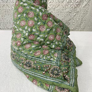 Może przedstawiać: Zwinięta zielona kołdra z kwiatowym wzorem. Wzór powtarza różowe i białe kwiaty z zielonymi liśćmi i łodygami. Obramowanie ma szczegółowy kwiatowy wzór. Kołdra wykonana jest z miękkiej, pikowanej tkaniny.