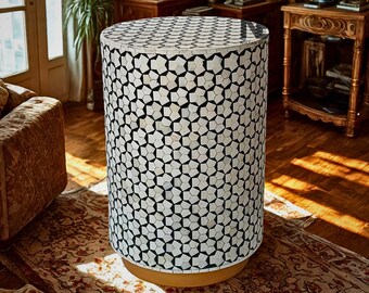 May include: A cylindrical side table with a geometric pattern. The table's surface is covered in a mosaic of white and black shapes, creating a star-like design. The base is a warm, light brown color. The table is in a room with hardwood floors.