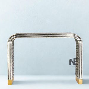 Könnte beinhalten: Eine dekorative Konsole mit geschwungenem Design. Der Tisch hat eine dunkelbraune Oberfläche mit einem weiß-schwarzen Punktmuster. Die Beine haben eine goldene Oberfläche. Der Tisch ist etwa 122 cm lang.