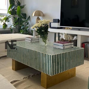 Puede incluir: Una mesa de centro rectangular con una tapa a rayas verdes y blancas y una base dorada. La mesa está en un salón, con un jarrón de flores blancas y libros encima. La mesa mide aproximadamente 122 cm de largo.
