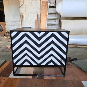 May include: A rectangular cabinet featuring two doors with a bold black and white chevron pattern. The cabinet has a dark brown top surface and is supported by a black metal frame base. The design consists of vertical zig-zag lines.