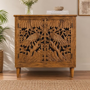 Puede incluir: Un gabinete rectangular de madera con dos puertas que presentan intrincados motivos tallados a mano de pavos reales y flores. El mueble se apoya sobre cuatro patas cónicas e incluye dos pequeños pomos circulares. La parte superior sostiene un cuenco y dos libros.