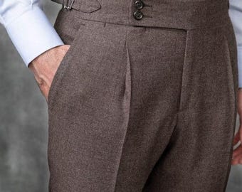 May include: Close-up of brown tailored trousers with a high waist and a buckle closure. The pants have a pleated front and a buttoned waistband. A light blue dress shirt is visible in the background.