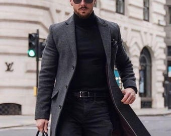 Puede incluir: Un hombre con un abrigo gris oscuro, un cuello alto negro y unos vaqueros negros camina por una calle de la ciudad. Lleva un maletín de cuero negro y gafas de sol y botas negras. El abrigo está abrochado y el atuendo es monocromático.
