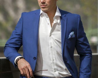 May include: A blue suit with a white dress shirt. The suit has a two-button closure and a pocket square. The person is wearing a watch and bracelet. The person is holding glasses.