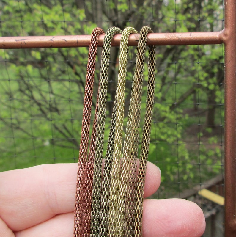Puede incluir: Cuatro collares de cadena en varios tonos de oro y cobre. Las cadenas est&aacute;n hechas de peque&ntilde;os cuadrados unidos y cuelgan de una barra de color cobre.
