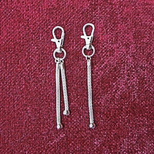 Puede incluir: Dos llaveros plateados con un borla de cadena. Cada llavero tiene un lazo plateado en la parte superior.