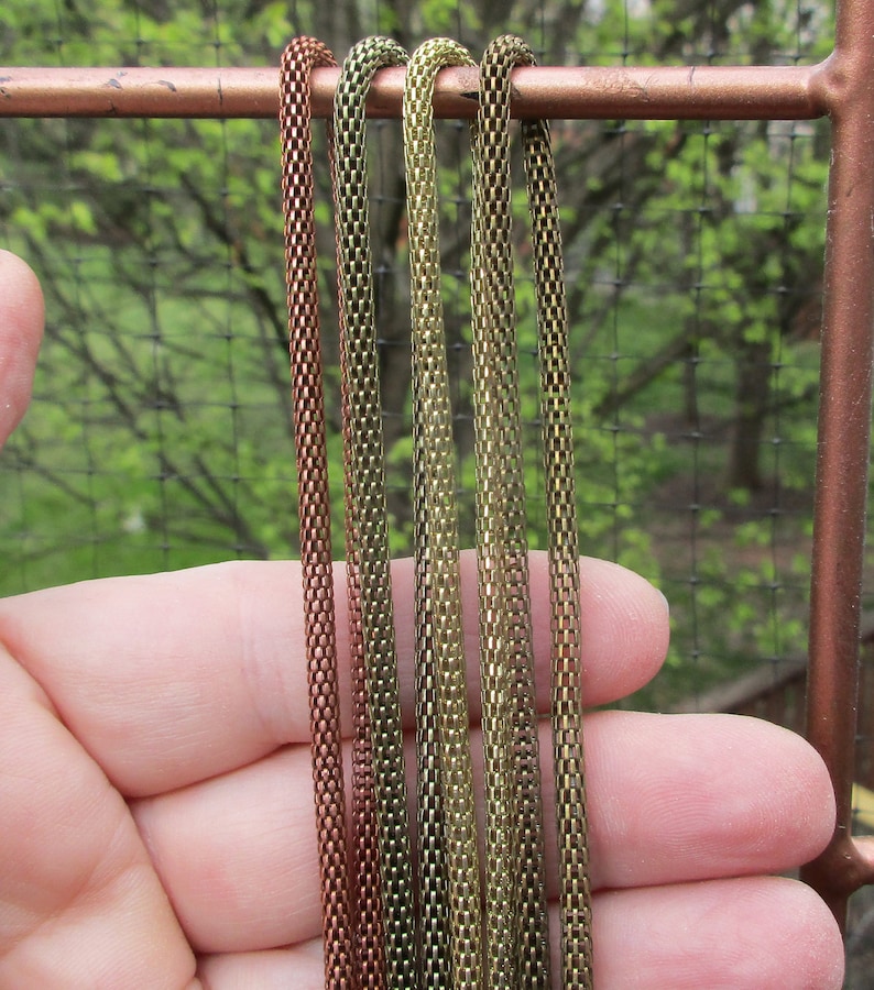 Puede incluir: Cinco estilos de cadena diferentes en tonos cobre y oro. Las cadenas est&aacute;n dispuestas verticalmente y se sostienen en una mano.
