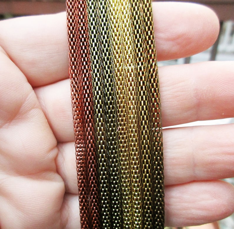 Puede incluir: Primer plano de una variedad de eslabones de cadena en diferentes colores, incluyendo cobre, oro y verde. Las cadenas est&aacute;n dispuestas en una l&iacute;nea vertical y se sostienen en una mano.